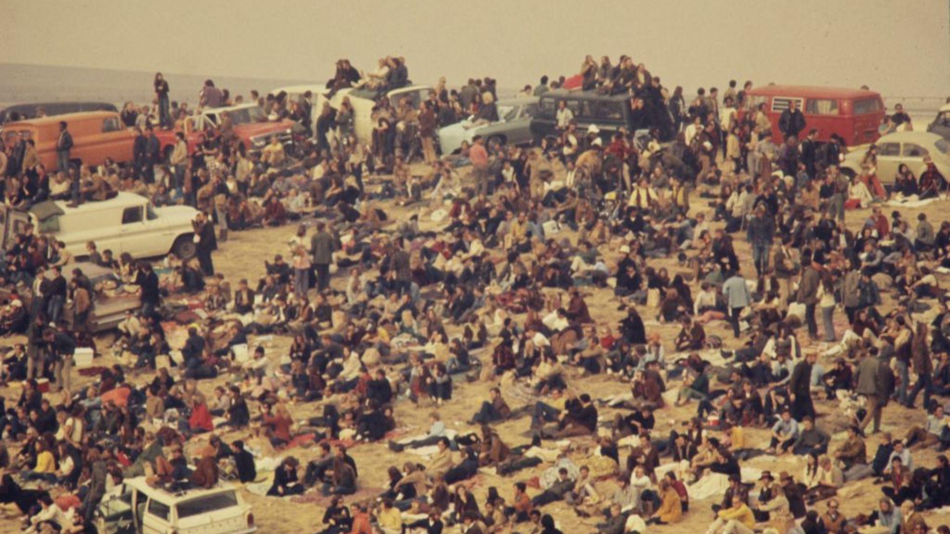 A large gathering of people sitting and standing around vehicles in a crowded parking area at an outdoor event. Many attendees are on the ground, while others are on top of vans and cars