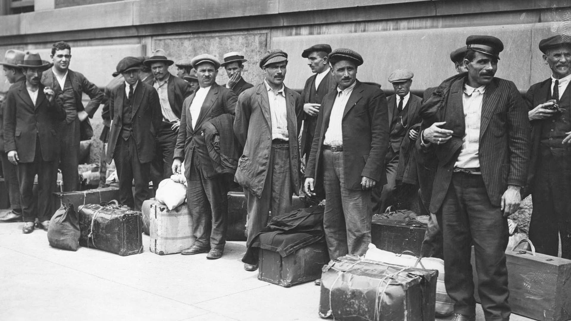 Black-and-white photo of Italian immigrants at Ellis Island in the early 1900s. A large line of male Italians stand in suits and hats with luggage at their feet.
