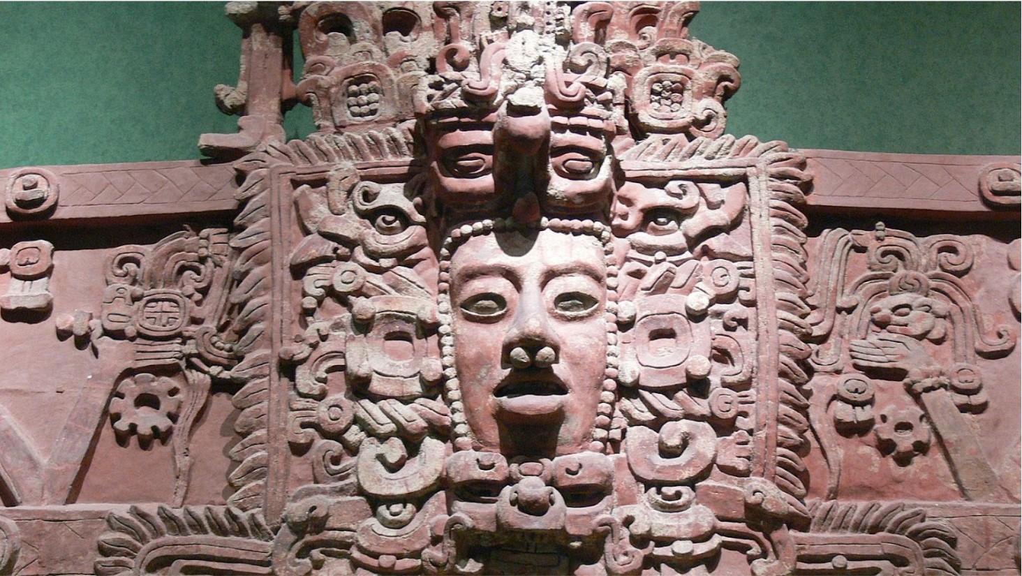 This is an image of an ornate Mesoamerican relief sculpture carved in stone, displaying a central face surrounded by intricate motifs and symbols that may represent a deity or royal figure