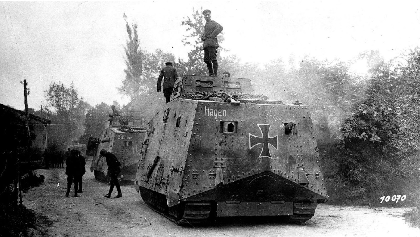 A black and white photograph of a World War I German armored vehicle, labeled 'Hagen,' on a village road. The tank has a prominent Iron Cross symbol on the side. Soldiers are visible around and on top of the vehicle