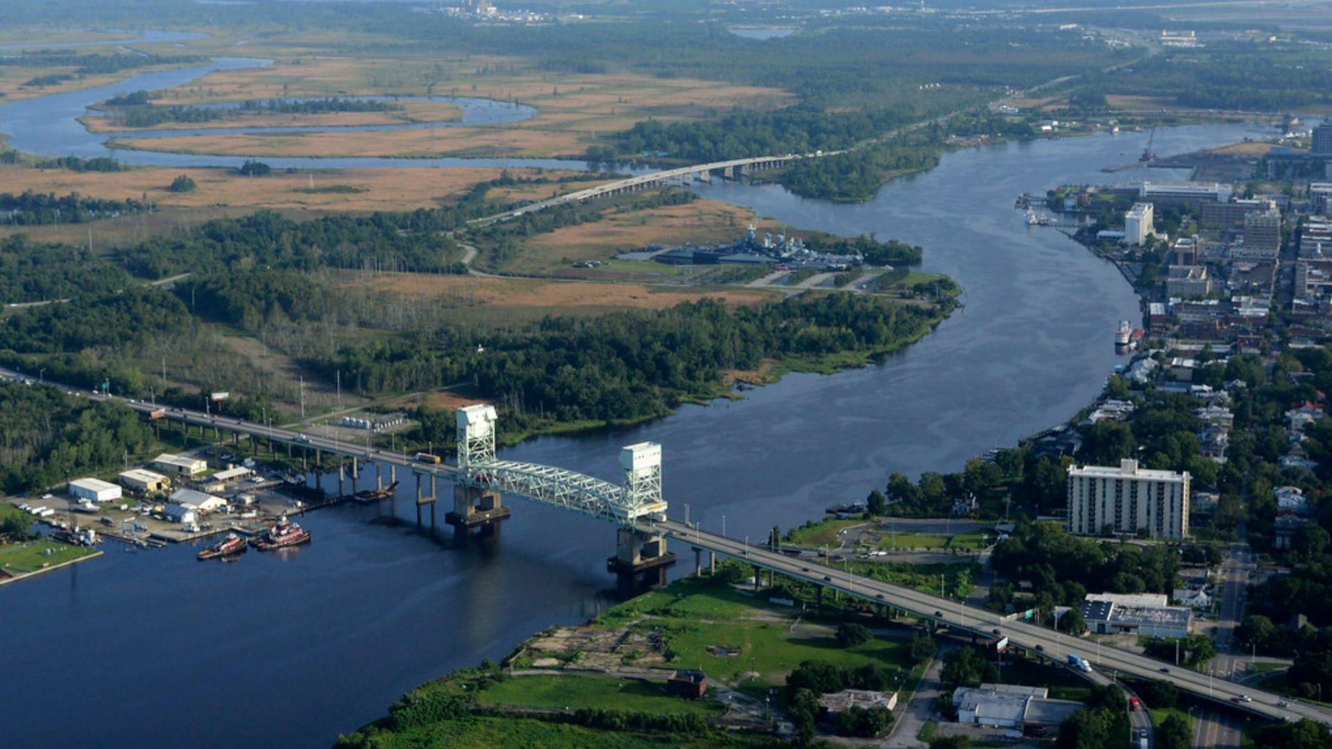 An aerial photograph captures a large drawbridge spanning across a wide river. The bridge features two prominent lift towers, allowing ships to pass beneath. On either side of the river, there are dense green patches of land with some areas appearing to be wetlands