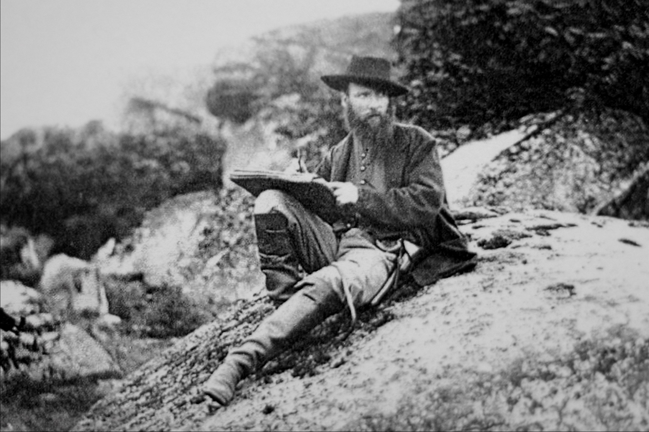 A man with a beard sitting on a hill drawing on a notepad