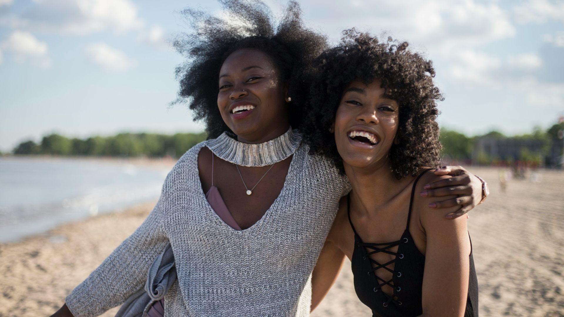 Two women smiling together with their arms around each other while at a beach.