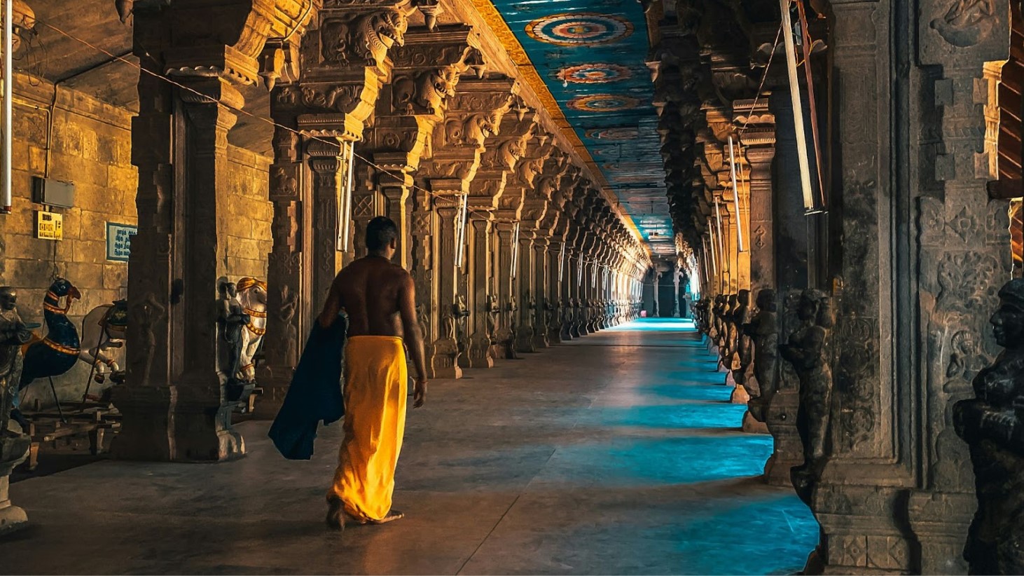 A lone individual draped in a traditional orange cloth walks through the grand corridor of an ancient temple, with by intricately carved stone pillars on either side