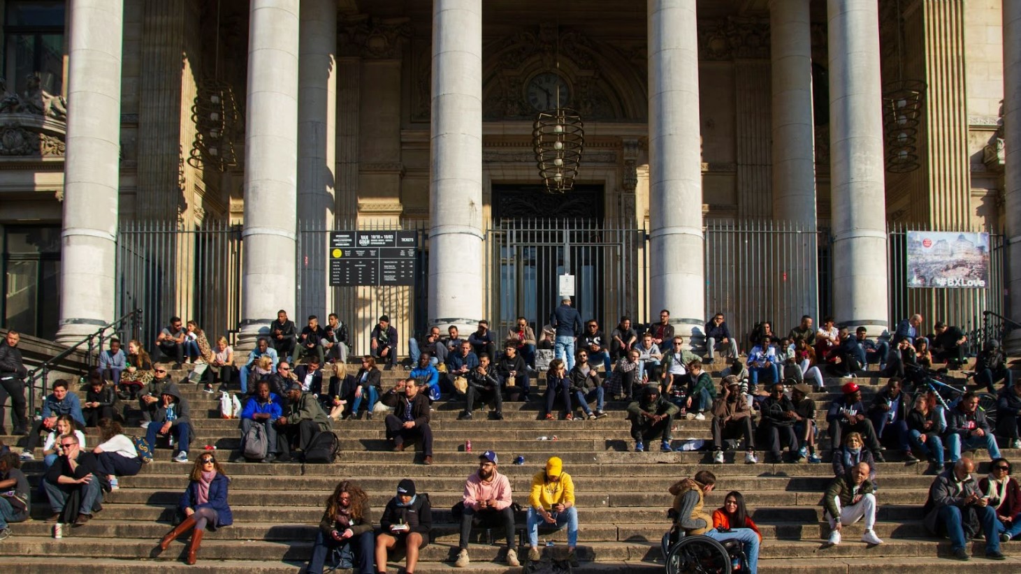A diverse group of people sitting and enjoying the sunshine on the wide steps in front of a grand building with tall pillars