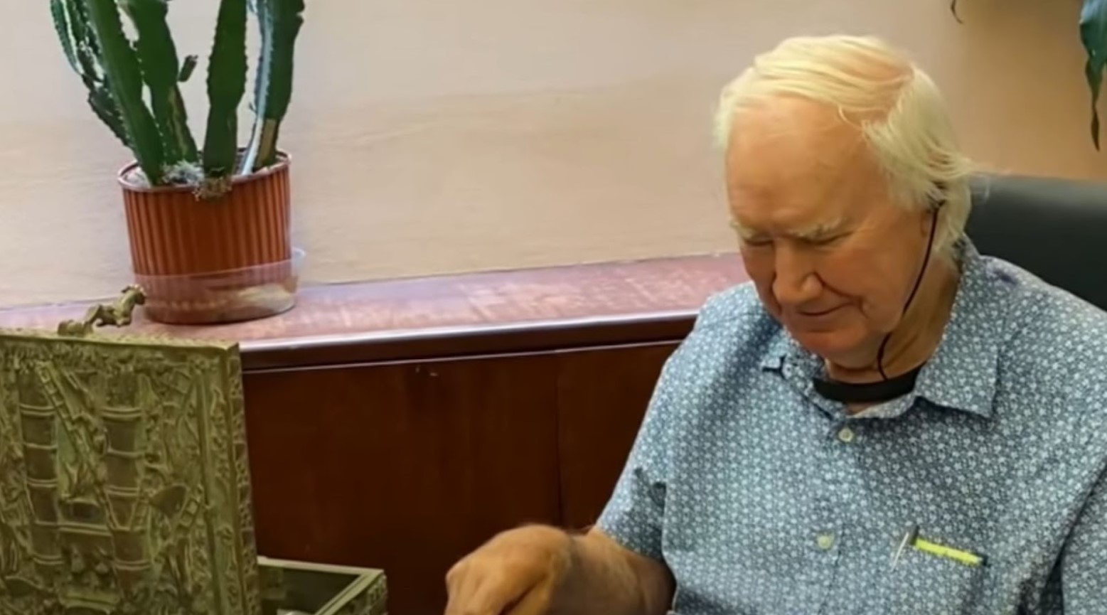 Forest Fenn sat with his treasure chest. There is a brown wooden side unit behind him with a cactus plant on top.