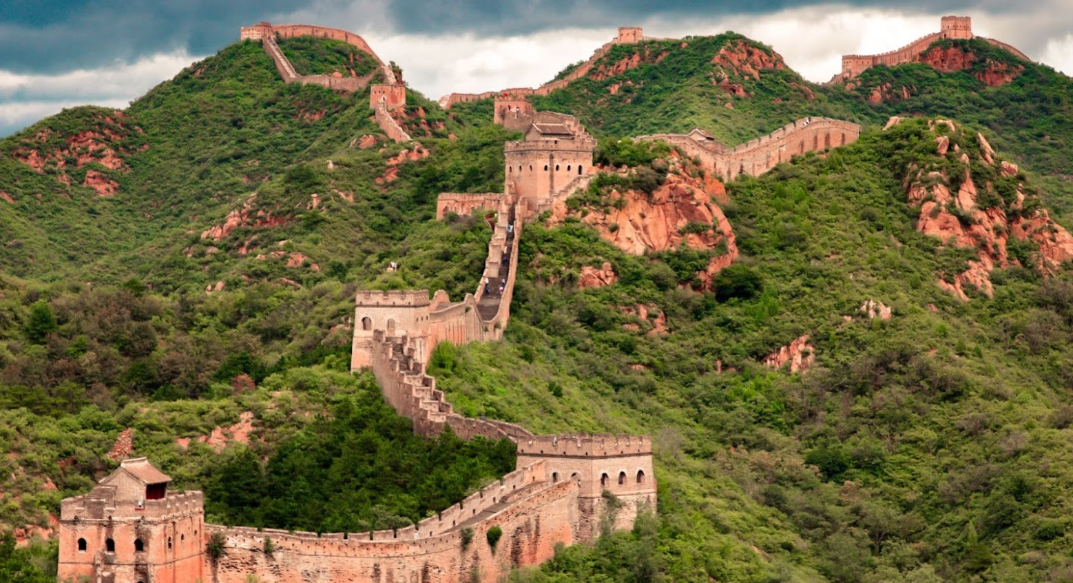 An image of the Great Wall of China. The wall goes up a hill and is surrounded by green trees and bushes.