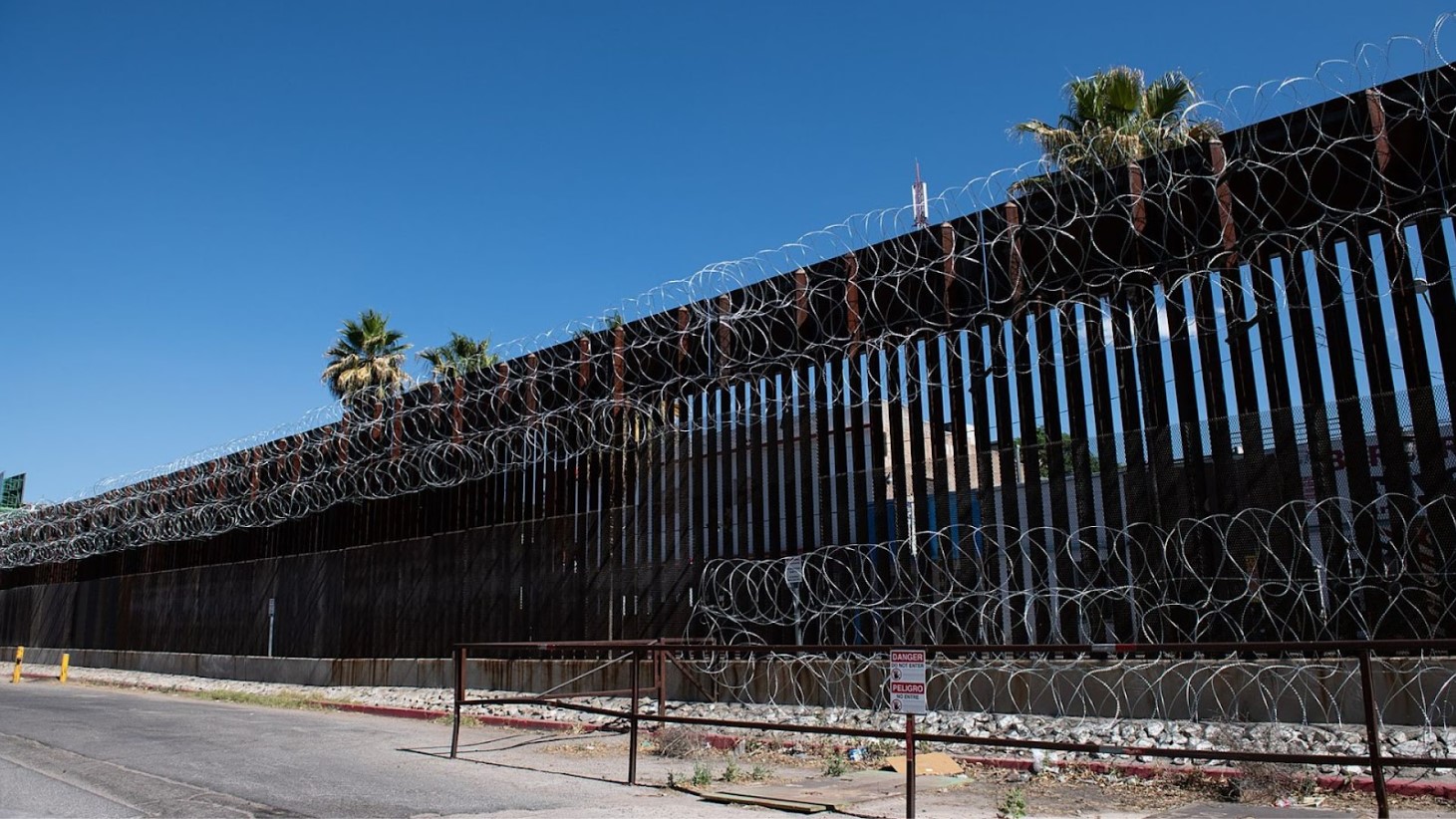 A tall border wall topped with multiple strands of barbed wire, with a clear blue sky above and a warning sign reading 'Danger' and 'Peligro' in the foreground