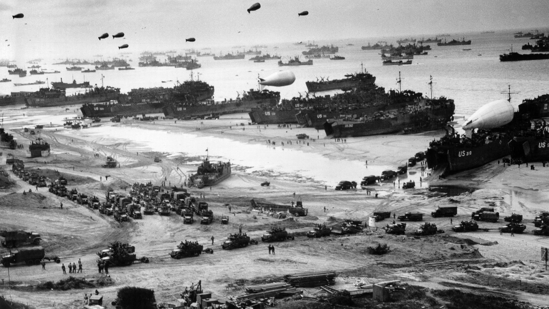 Black-and-white photo of landing ships putting cargo ashore on Omaha Beach in Normandy, France at low tide during the early days of the Allied invasion, mid-June 1944.