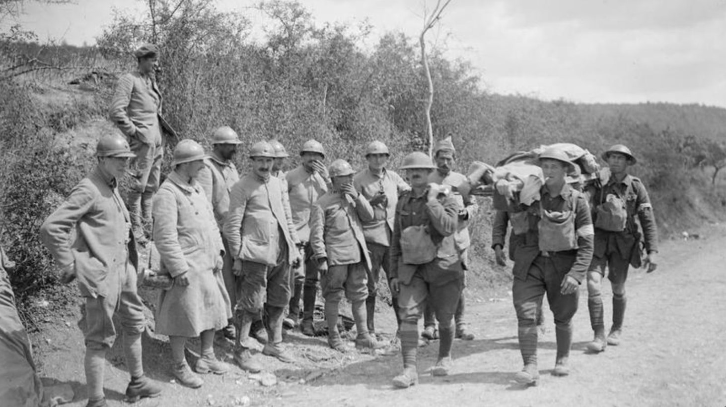 A black and white photograph of World War I soldiers standing and walking along a dusty roadside. Four soldiers are carrying a wounded man on a stretcher