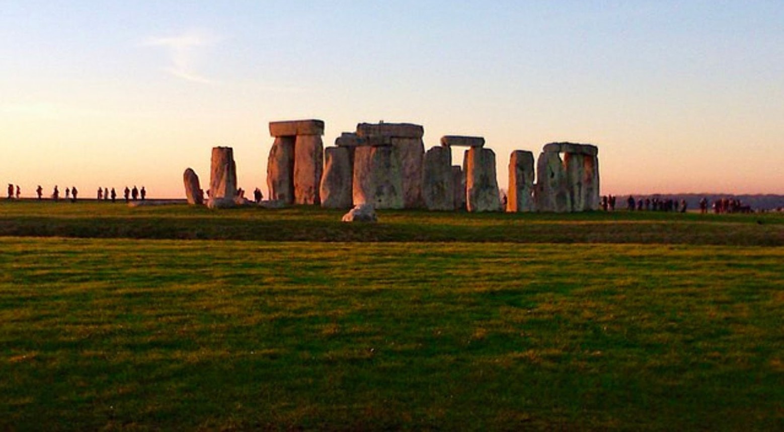 Stonehenge at sunset. There are visitors on either side of the stones to watch it.