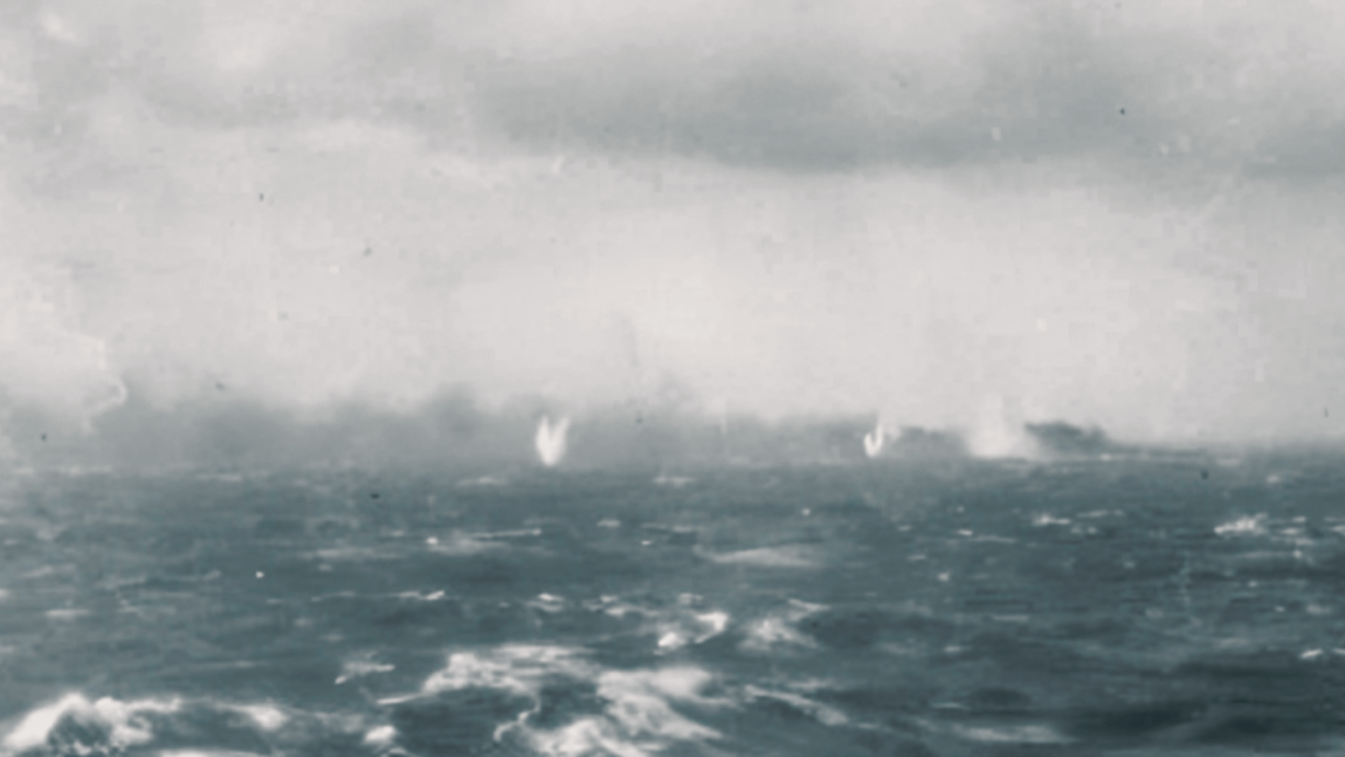 A blurry photo of the Bismarck burning and sinking while surrounded by splashes from shelling.