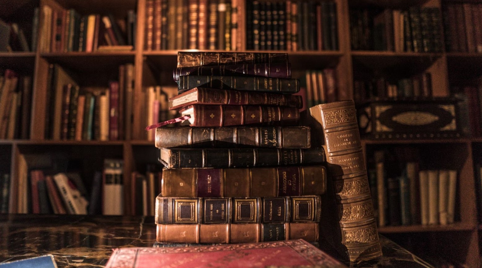 A pile of old books stacked on top of each other, with one book leaning against the pile. Behind the pile of books is a bookshelf full of books.