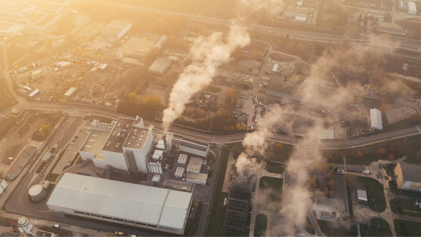 An aerial view of an industrial area during the golden hour of dusk. The area is characterized by multiple buildings with large smokestacks releasing plumes of smoke into the air