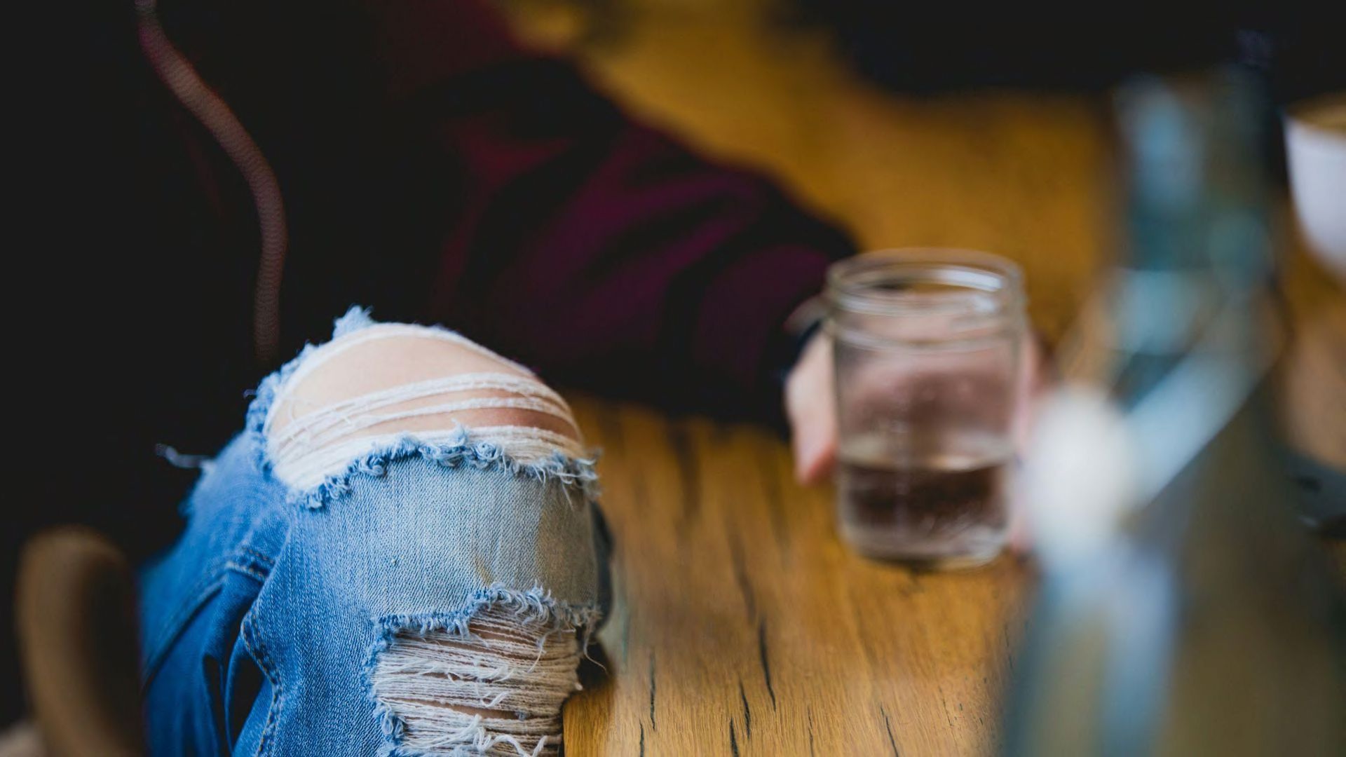 A person sitting by a wooden table wearing ripped jeans and holding a glass.