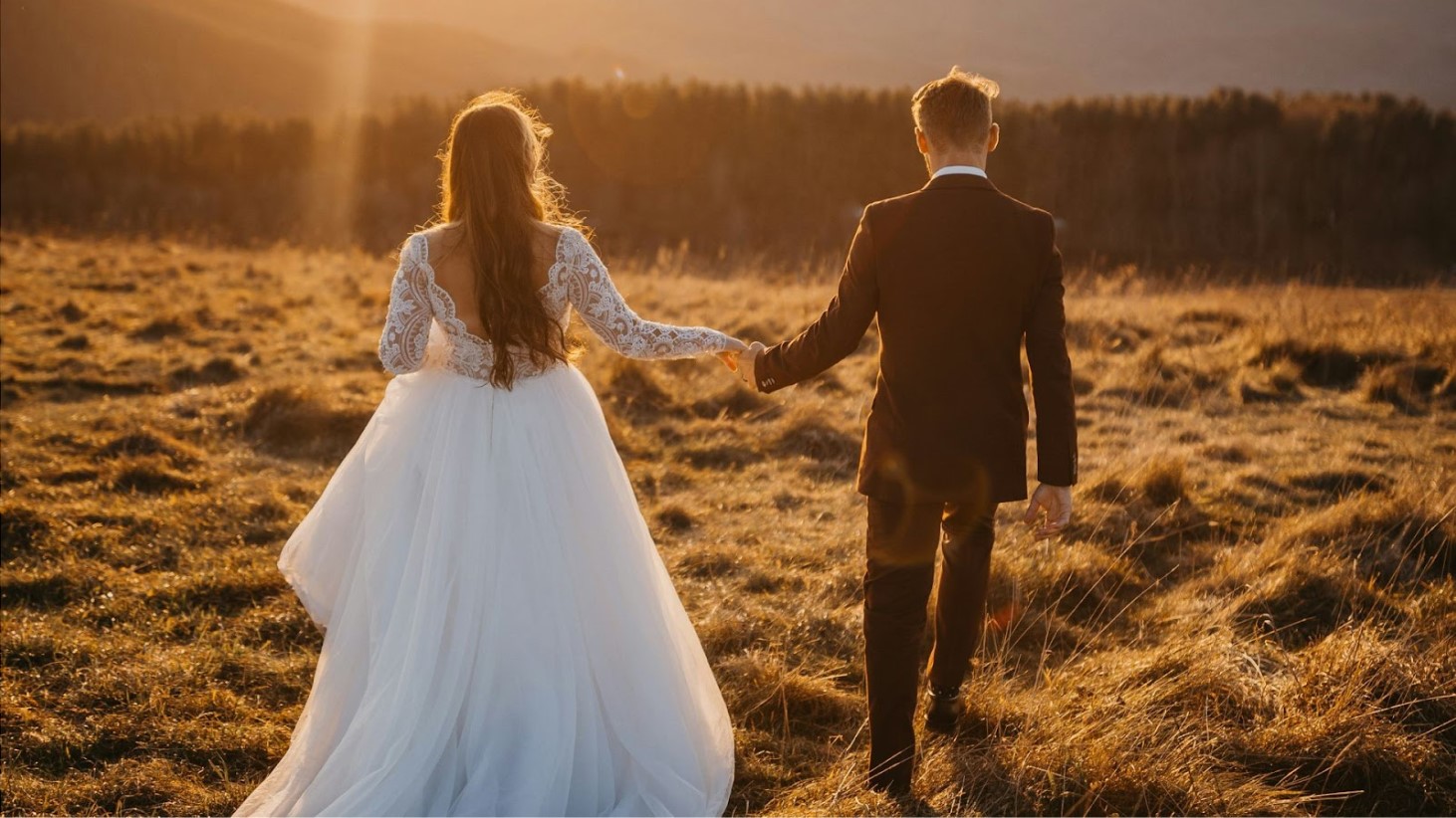 A bride and groom, viewed from behind, holding hands and walking through a field with dry grass at sunset