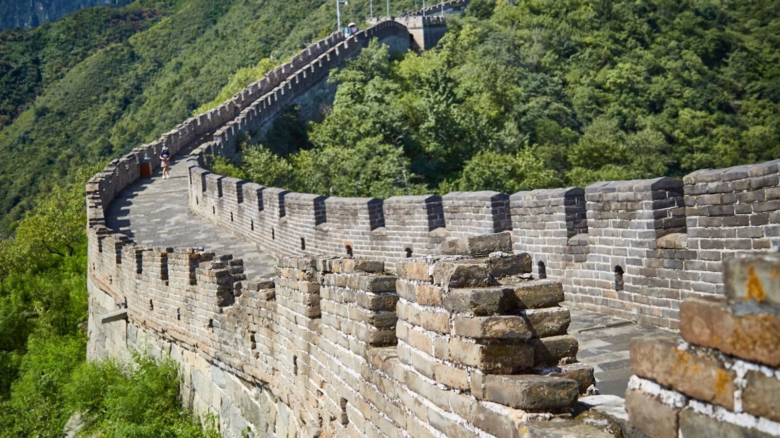 A close-up image of the Great Wall of China. The wall curves around and goes up and a couple of people are walking up it.