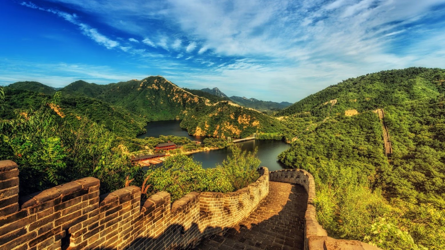 The Great Wall of China during summer. The photo is taken from some steps going down and is surrounded by green trees and bushes.