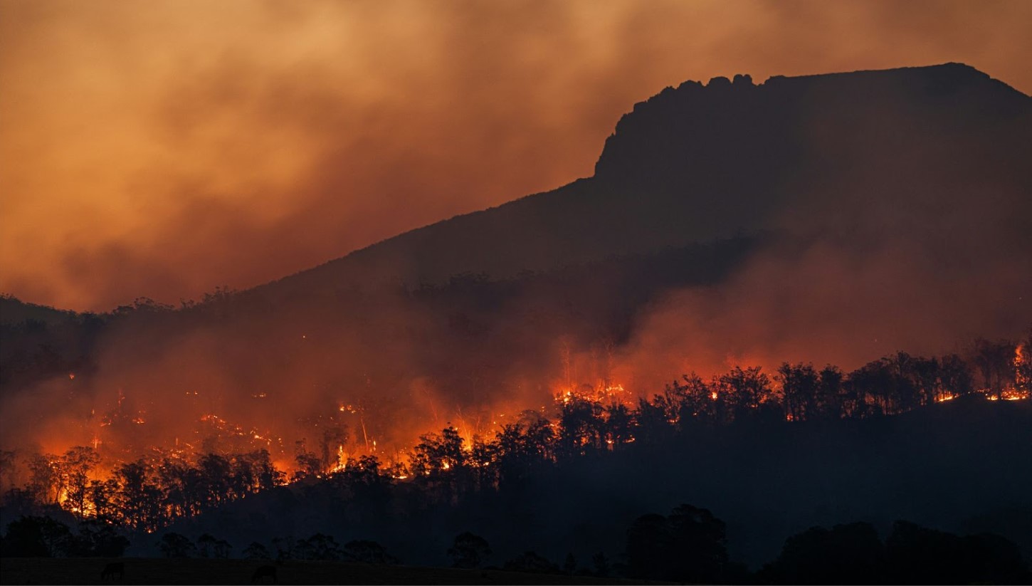The image shows a dramatic scene of a wildfire in a mountainous region. The lower slopes of the hill are engulfed in flames and smoke, with the fire casting an intense orange glow against the smoke-filled sky