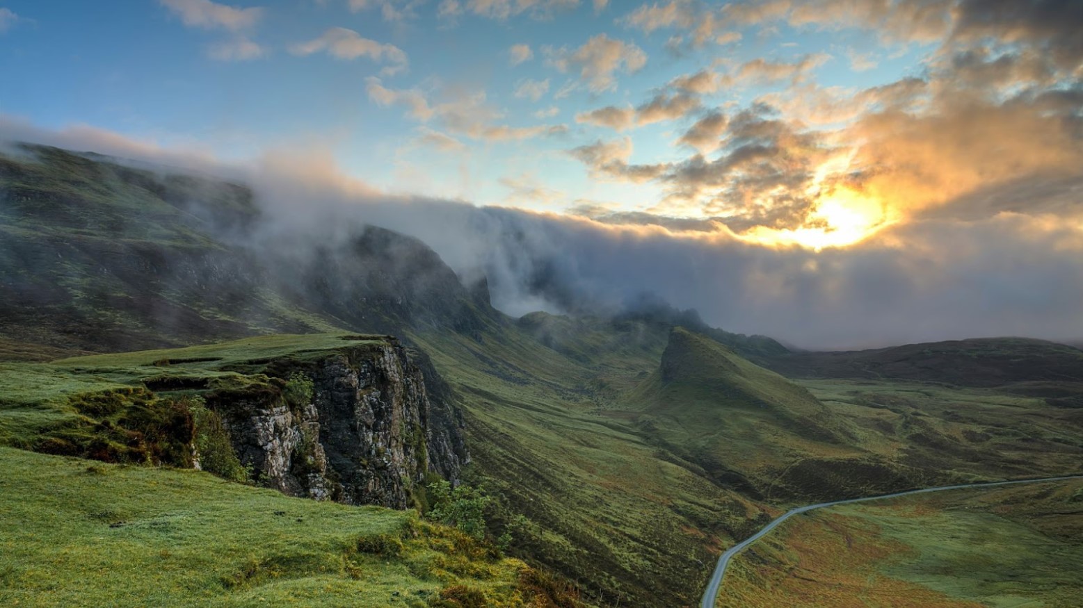 Green cliffs, hills, and pastures underneath a cloudy blue sky in the daytime.