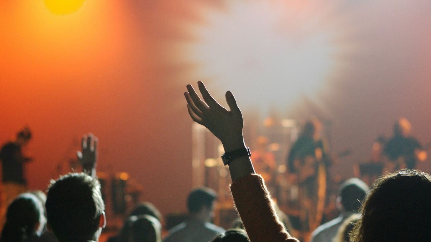 A silhouette of a person's raised hand in a crowd at a concert, with the stage in the background illuminated by a warm, orange light