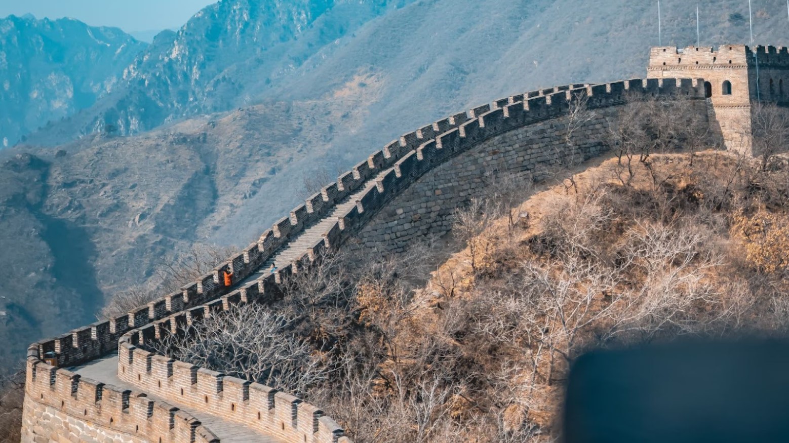 The Great Wall of China. The wall is going up and there is a person in a red jacket about to walk up the steps of the wall.