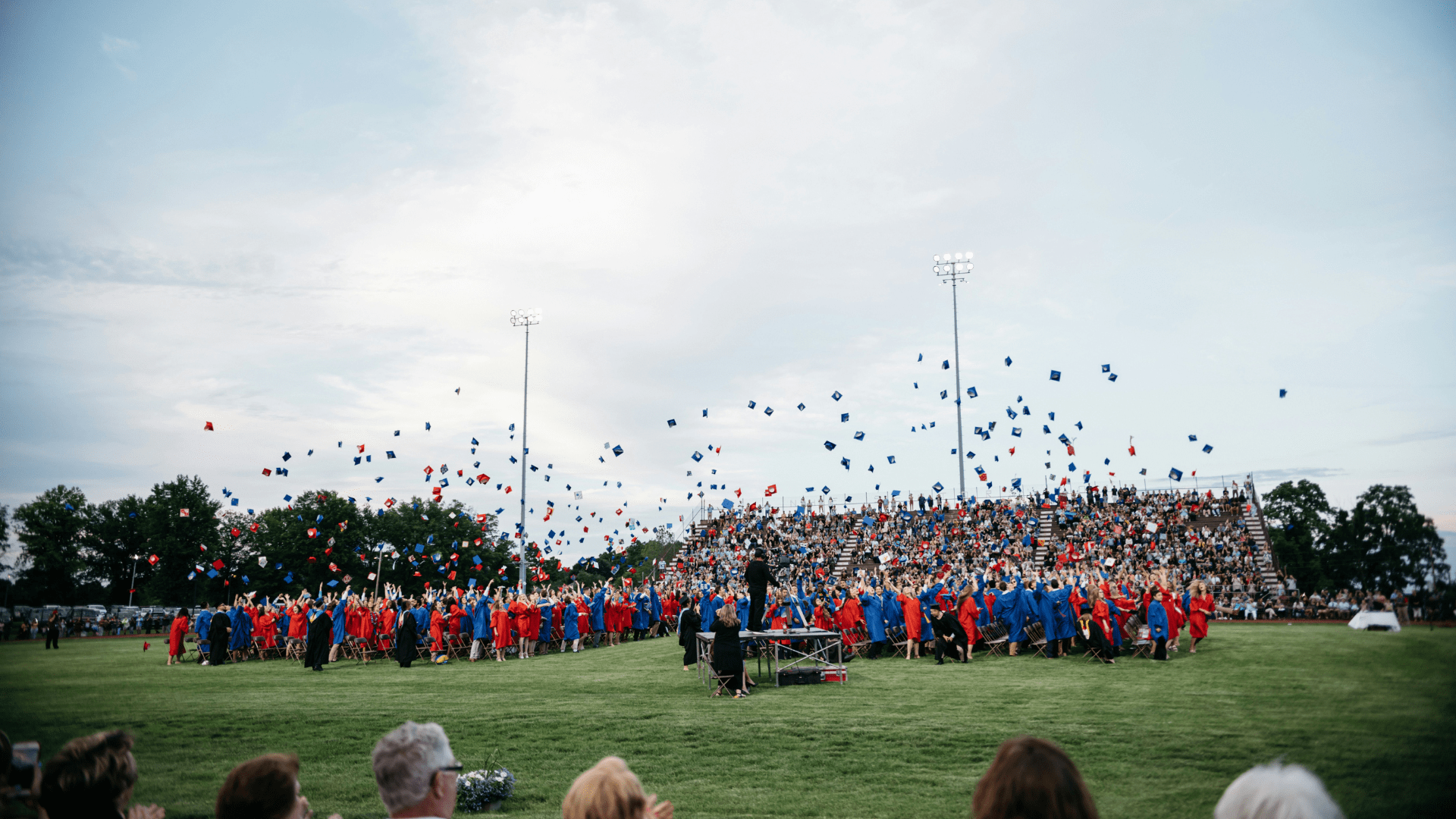 A field full of students graduating from high school wearing red and blue robes and hats. They’ve just thrown their hats into the air.