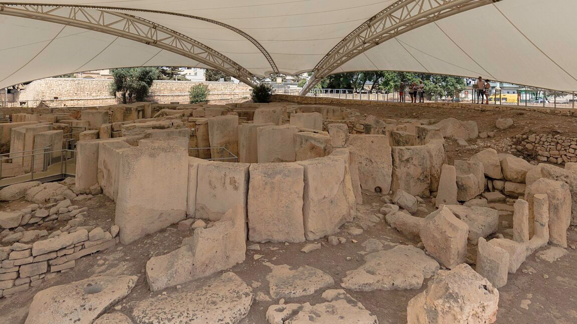 An aerial view of the megalithic temple ruins in Malta, covered by a protective white canopy. The complex consists of large limestone blocks, some standing upright and others toppled over, forming ancient structures and doorways. Walkways for visitors are visible around the site