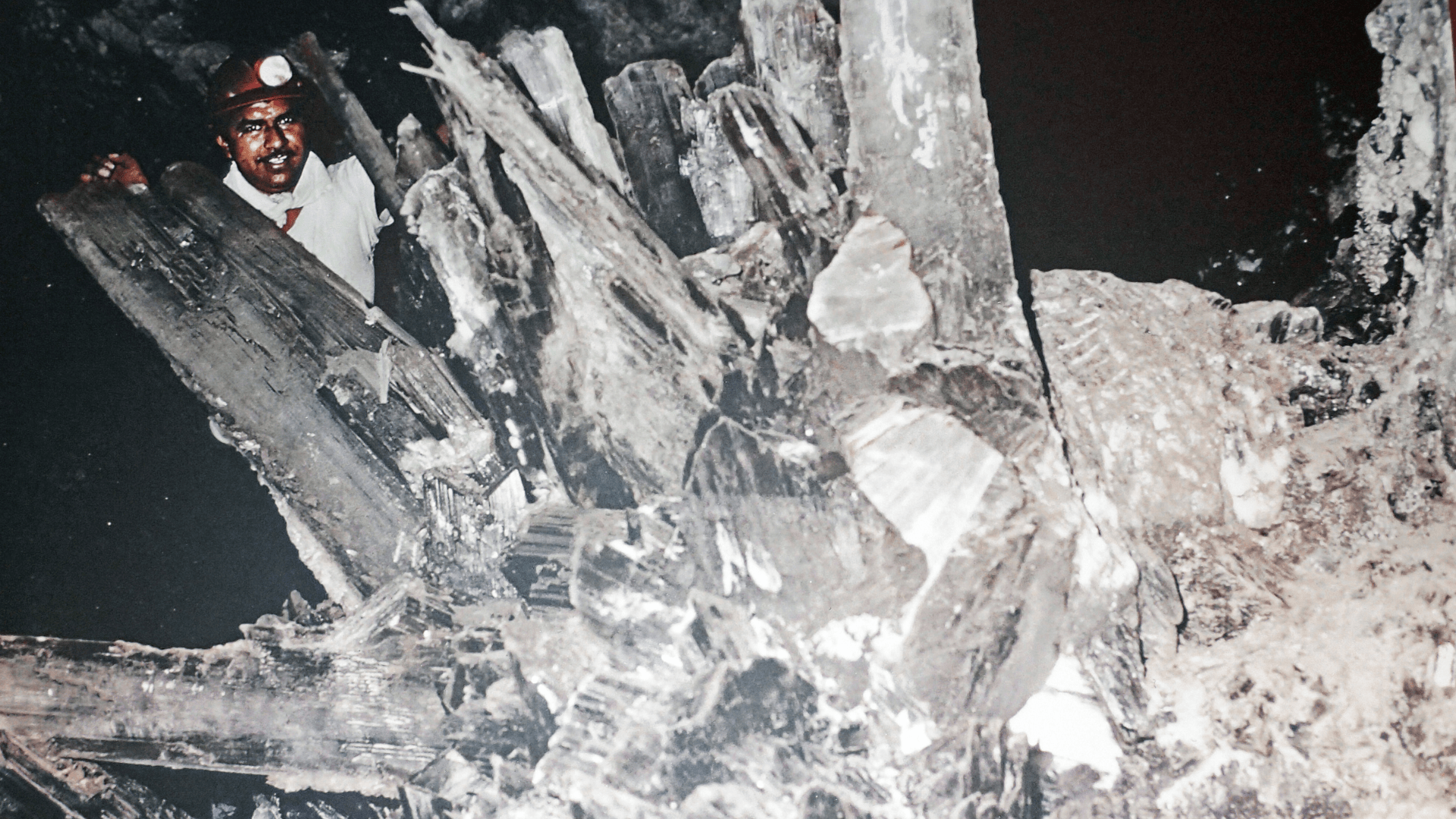A man with a helmet and light stands with large gypsum crystals in the Naica Crystal Mines.