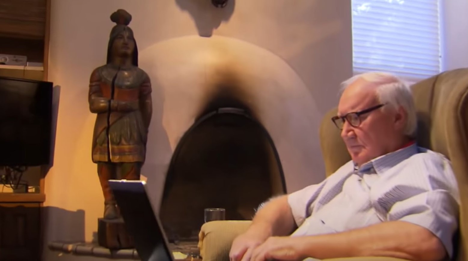 Forest Fenn sat in an armchair looking at his laptop. There is a bronze statue to the side of him.