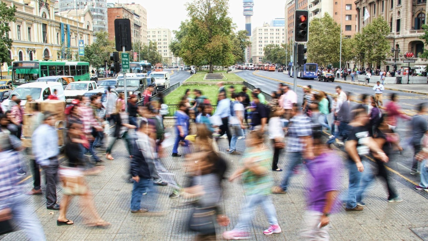 A blurred motion photograph capturing a busy city street crossing during rush hour. Scores of pedestrians are crossing in various directions, with their movements appearing in a blur