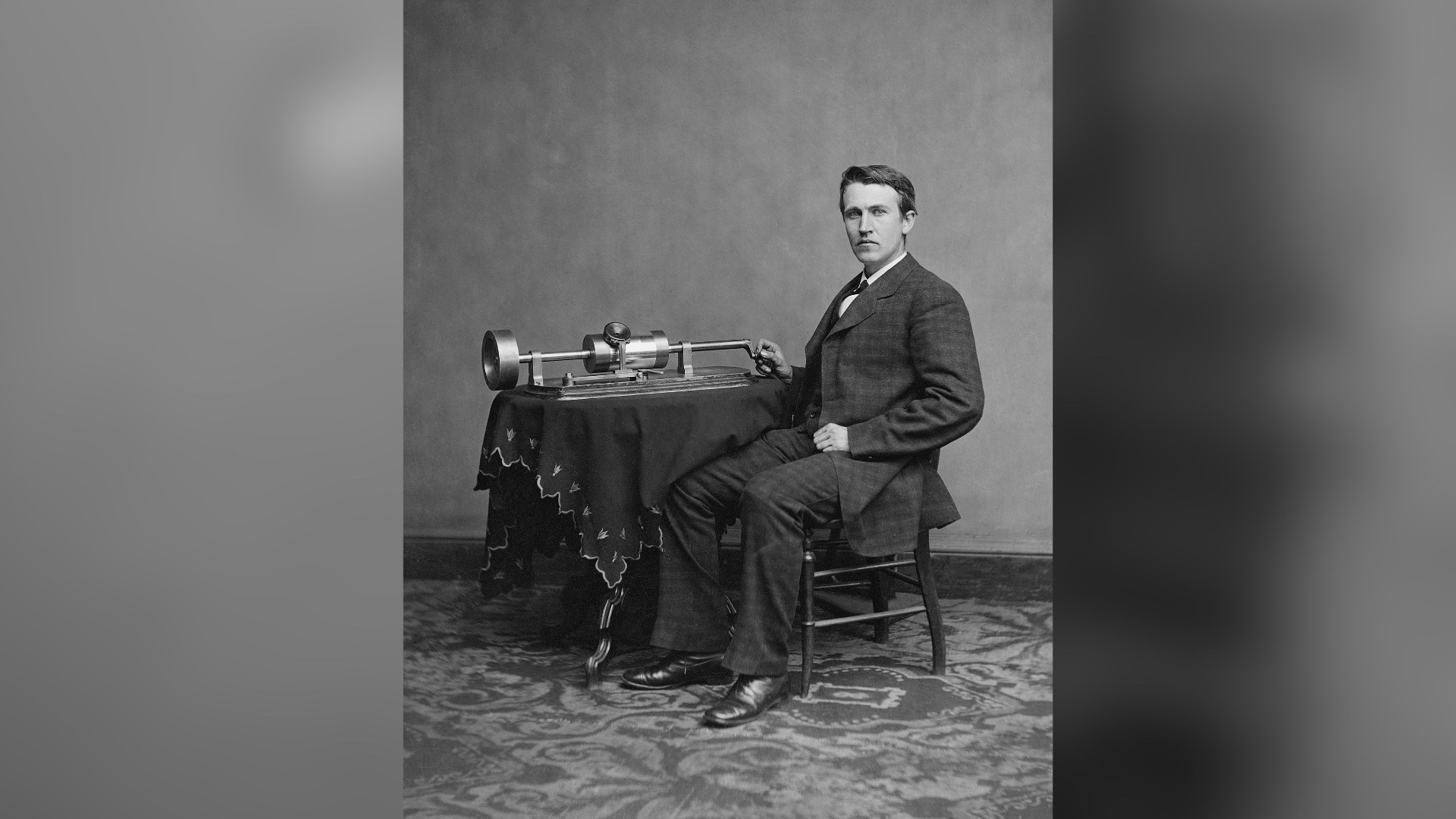 Black-and-white photo of Thomas Edison sitting at a phonograph wearing a suit.