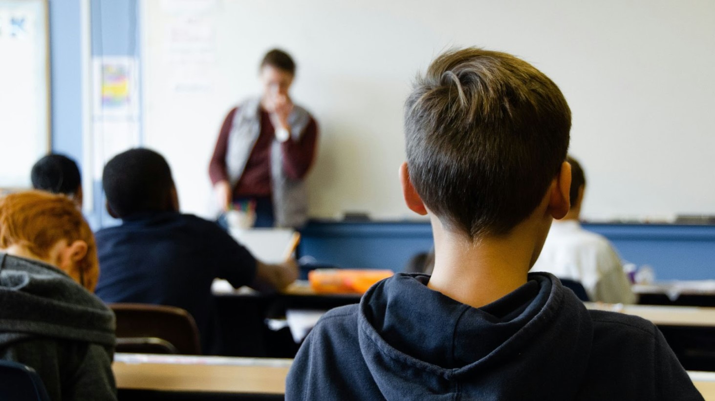 A classroom scene from the perspective of a student sitting at a desk, with the back of a student's head in the foreground, other students in the middle ground, and a teacher standing by a desk in the background