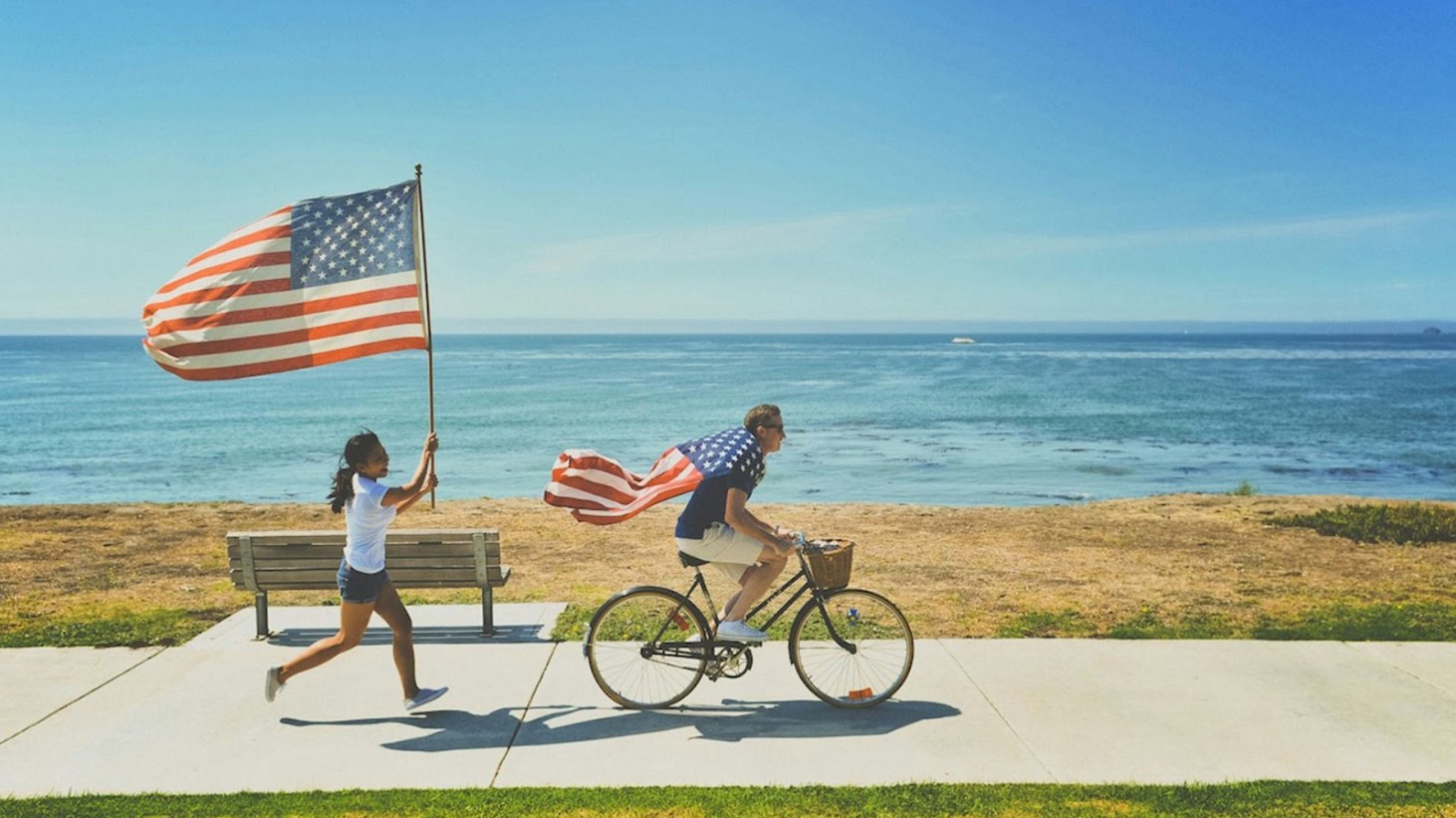 A person riding a bicycle along a coastal pathway with an American flag draped over the back, followed by a young girl running with a smaller American flag, both against a backdrop of the ocean and clear blue sky