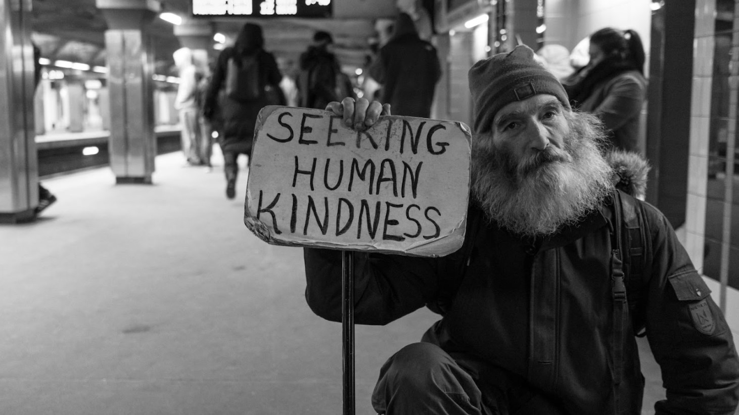 A black and white image capturing a bearded man holding a sign that reads "SEEKING HUMAN KINDNESS" in bold, hand-written letters