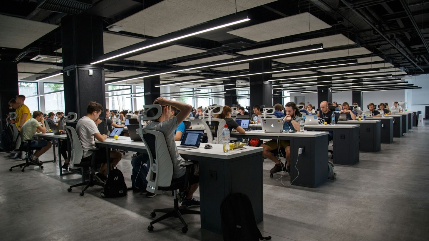 A wide view of a modern office space filled with people working at desks with computers, set against an industrial-style ceiling with hanging lights