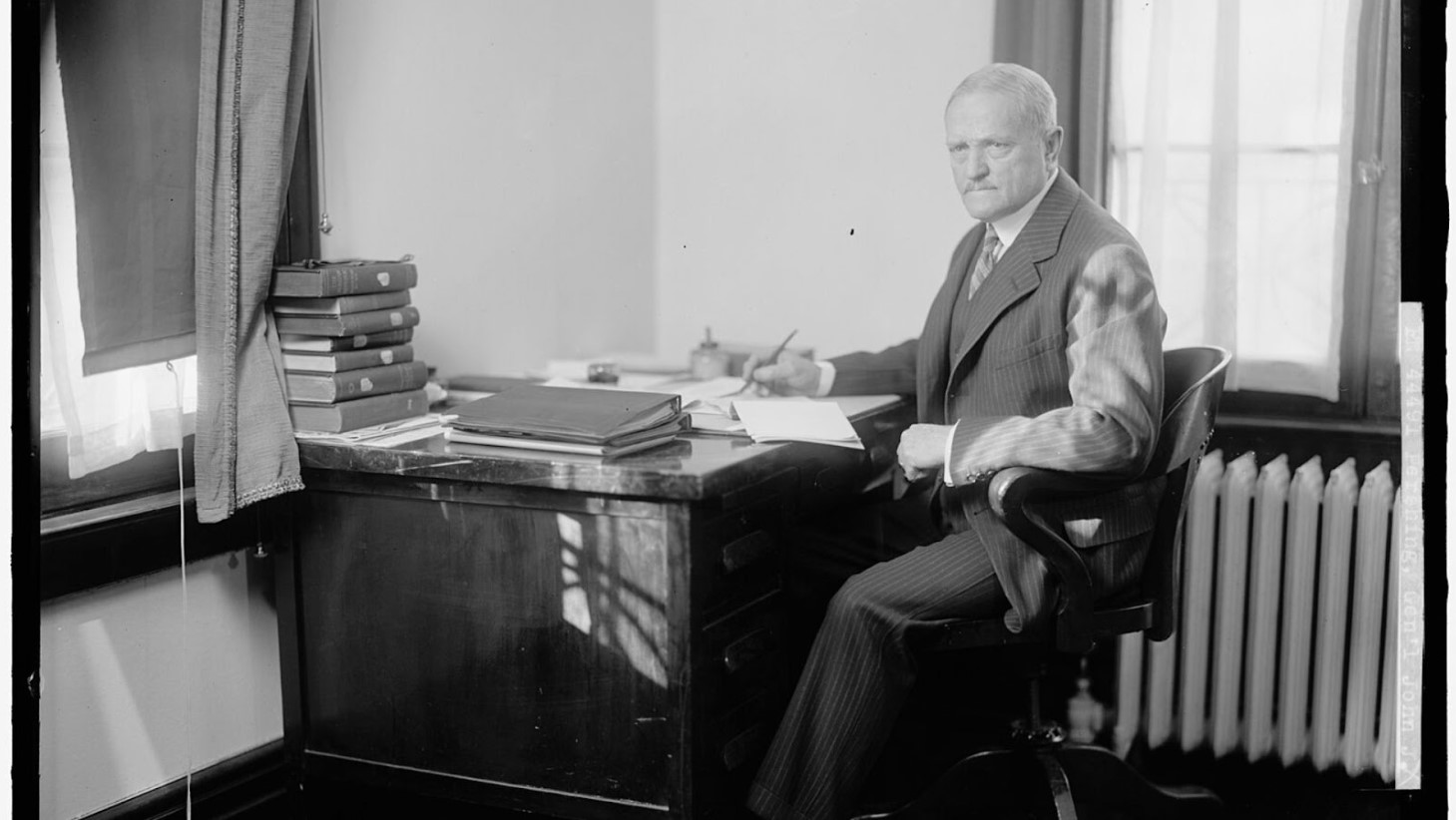 Black and white photo of an older man seated at a desk, looking directly at the camera. He is dressed in a pinstripe suit, with a pen in hand over a pile of papers. A stack of books sits on a window ledge behind him