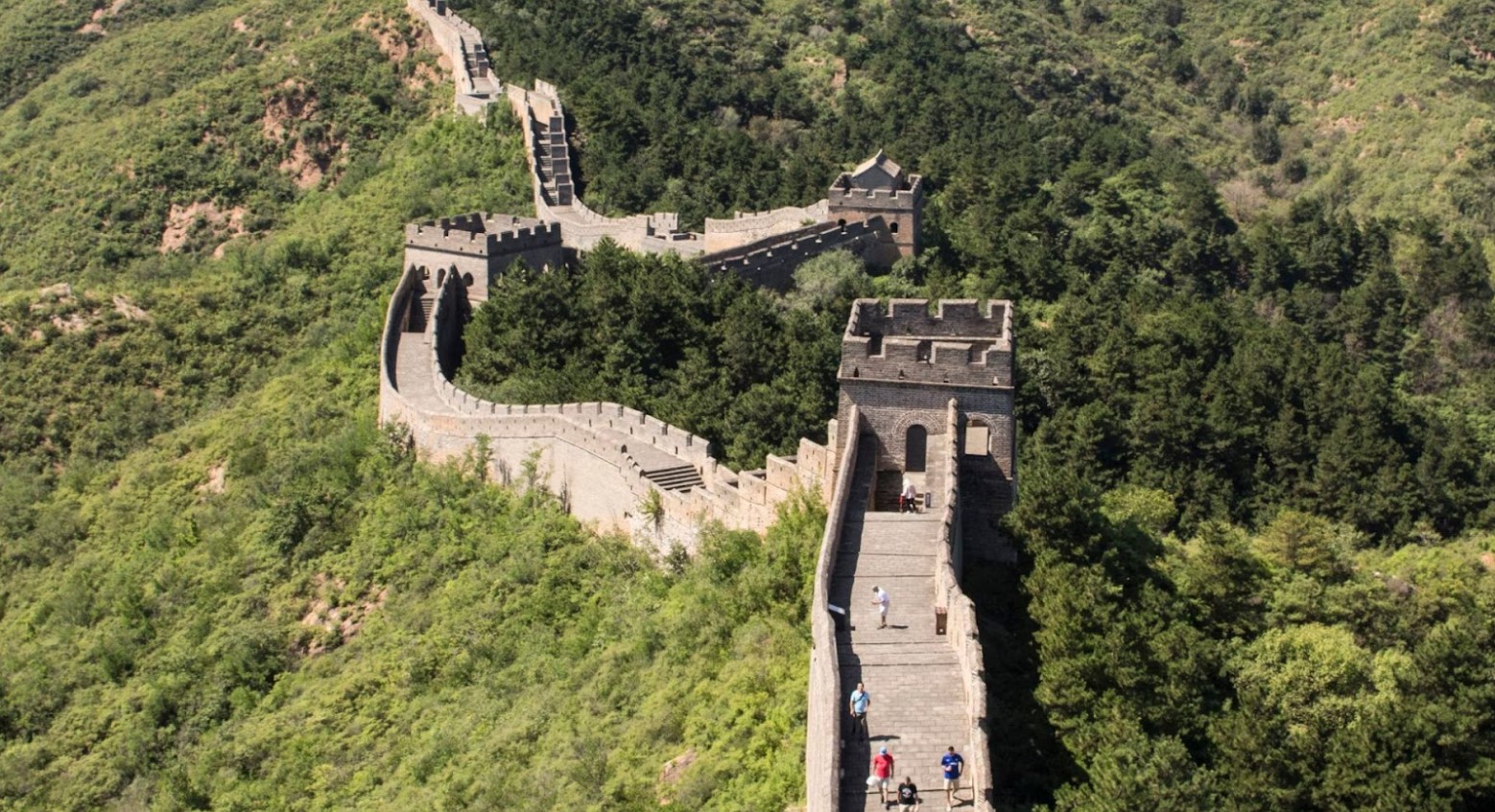 An aerial view of the Great Wall of China. Some people can be seen walking along the wall and there are green bushes and trees on either side of it.