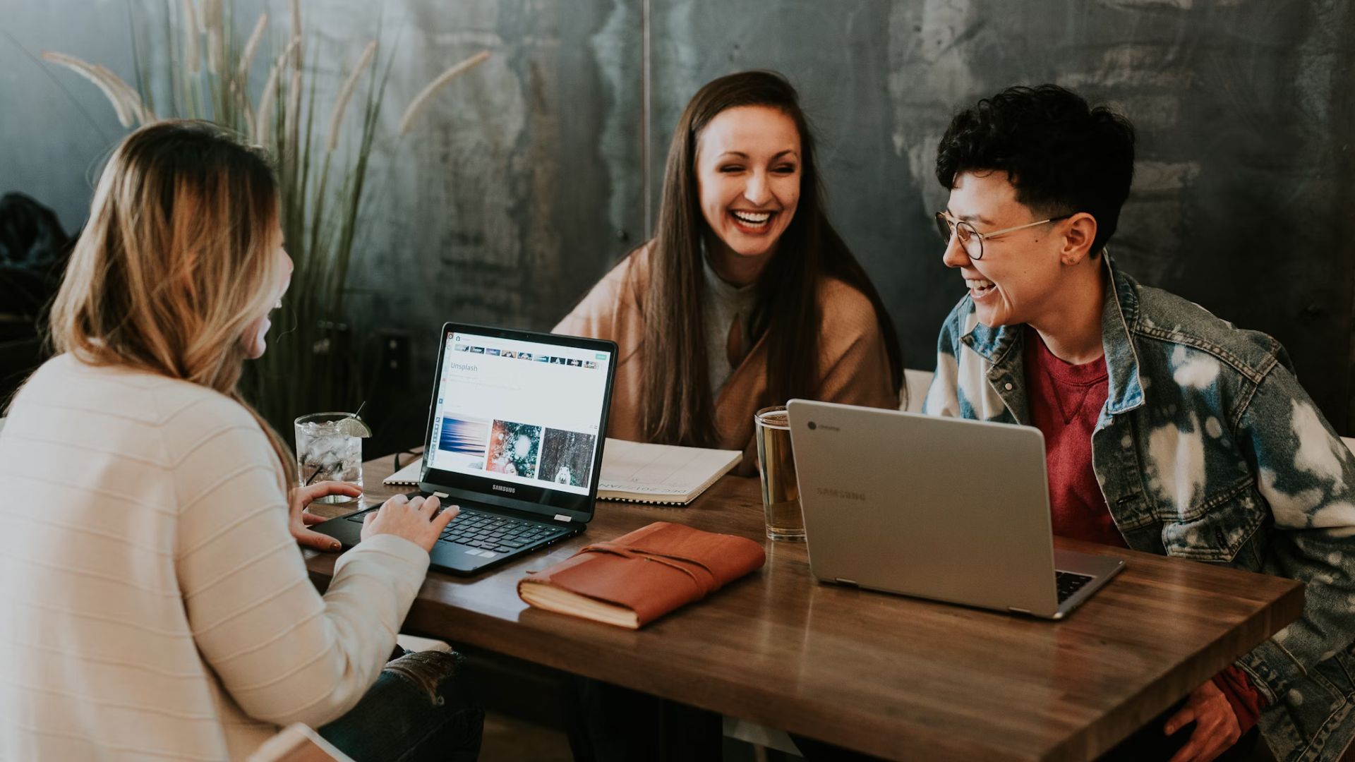 Three people sat around a wooden table working. There are each using a laptop and are laughing and smiling.