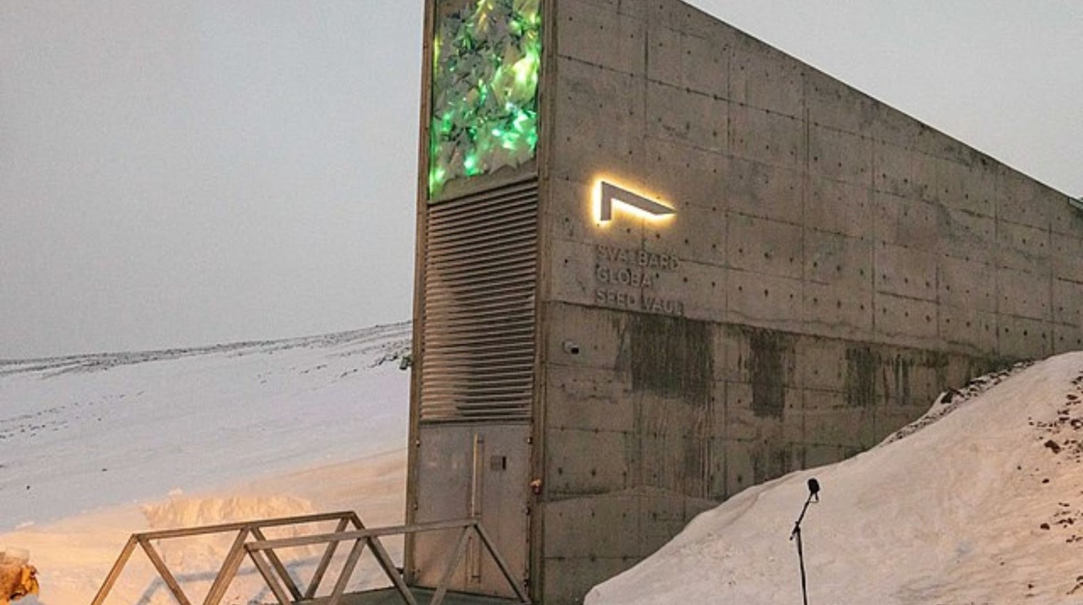 The outside of the seed vault in Norway. The vault is surrounded by snow. The vault is grey with double doors to get into it. There is a sign for the seed vault on the side of the vault, which looks like the number ‘7’ on its side, which is lit up around the edges.