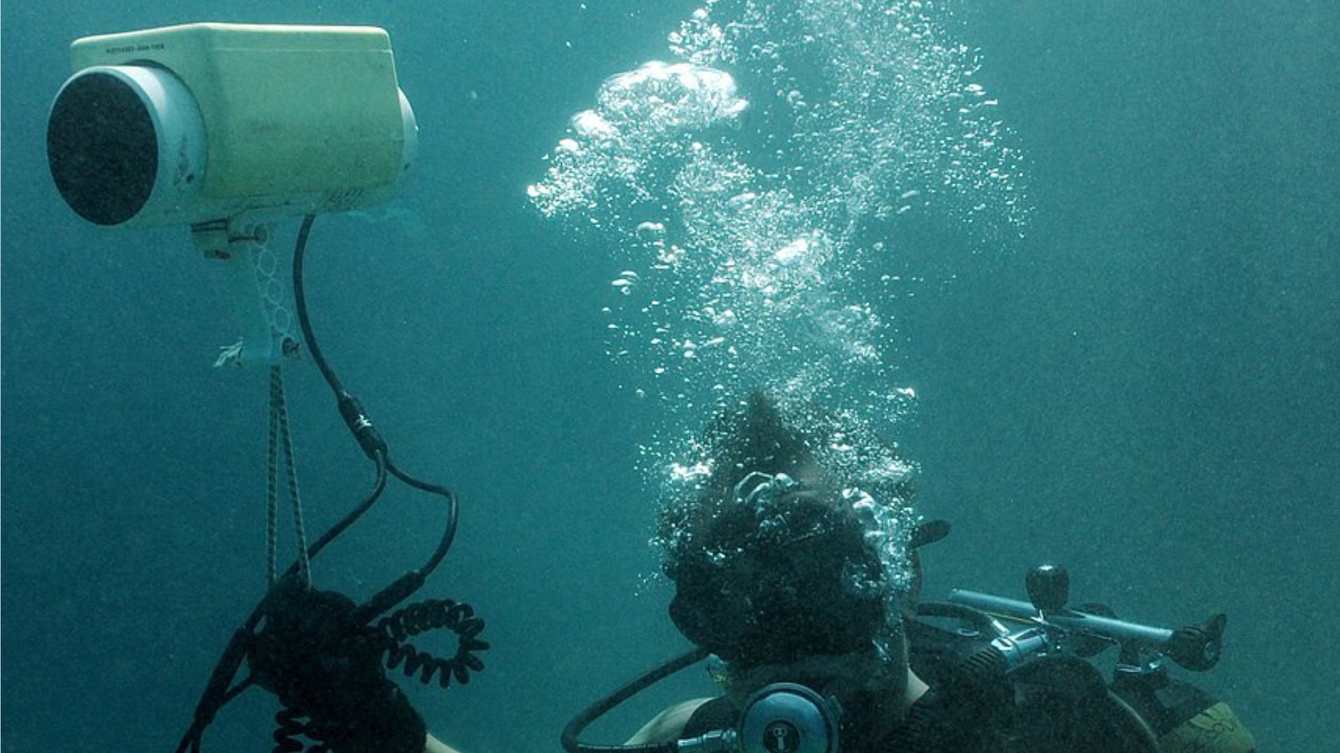 An underwater scene capturing a scuba diver next to a subsea sonar system mounted on a tripod. The diver, clad in dive gear with a mask and regulator, is releasing a stream of air bubbles that are ascending towards the surface
