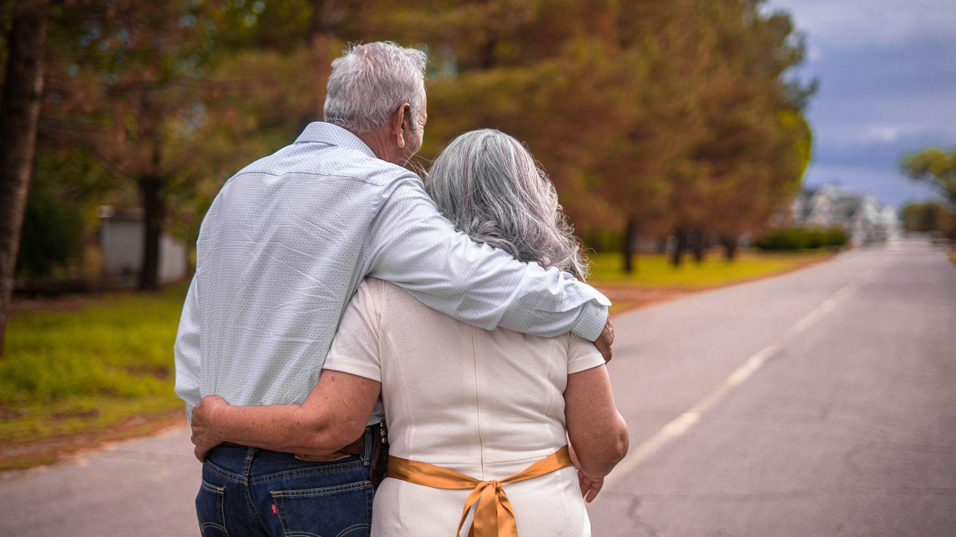 An old couple walking with their arms around each other in the daytime.