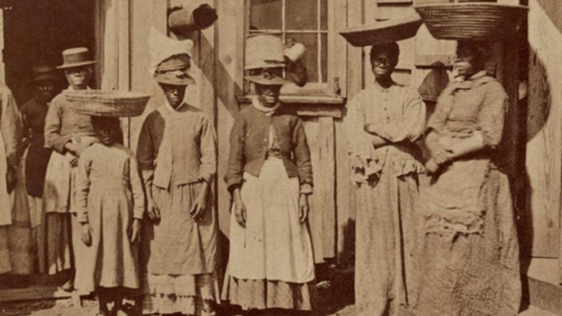 A sepia-toned photograph showing a group of African American women and children standing outside a wooden building. The women are dressed in long dresses and aprons, and most are wearing hats; two are balancing baskets on their heads. A child stands in the foreground