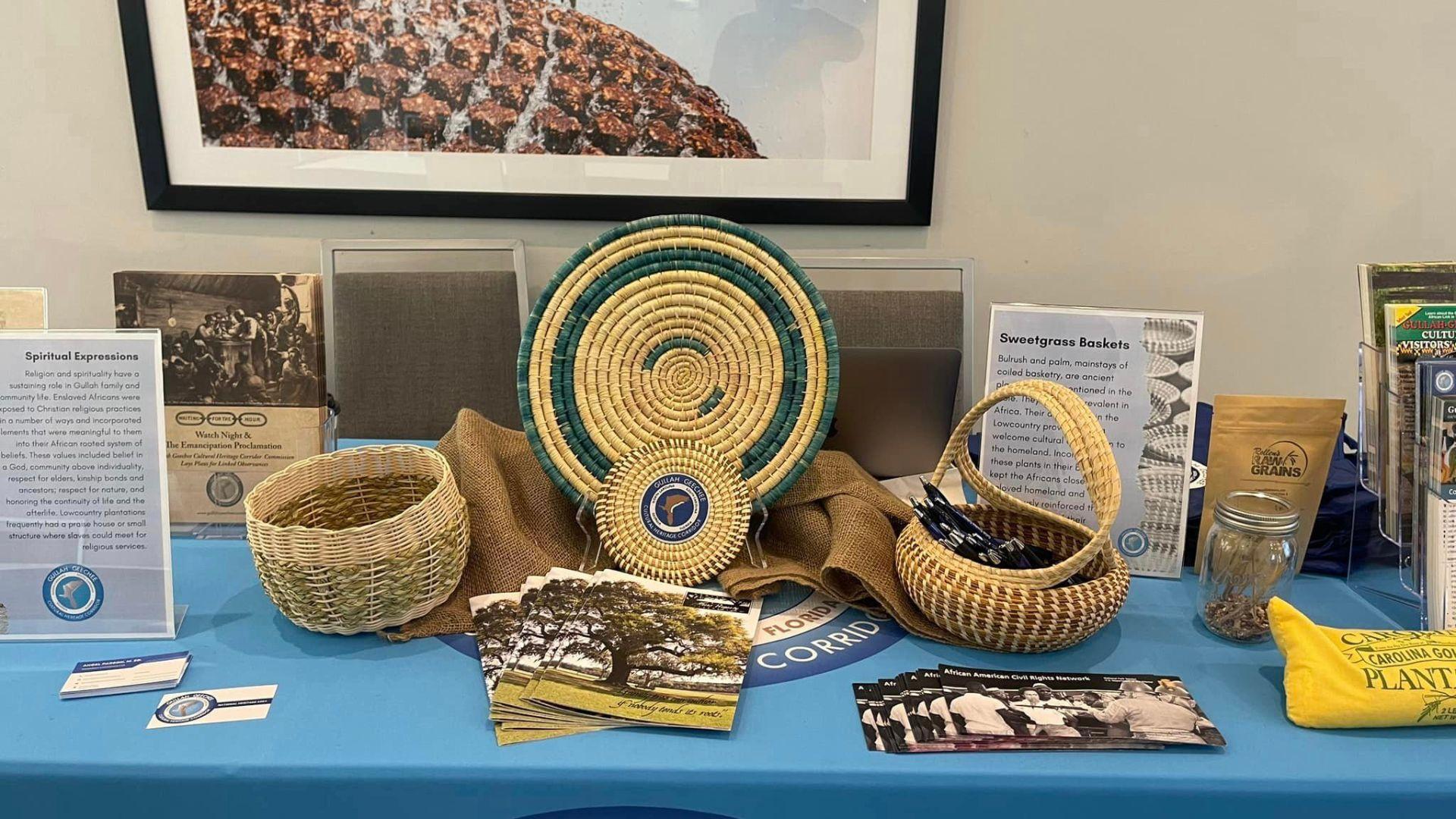 A table is set up with an exhibition of Gullah Geechee cultural artifacts and informational materials. In the center stands a large, intricately woven sweetgrass basket in green and natural hues. Flanking it are smaller baskets, brochures, and informational cards