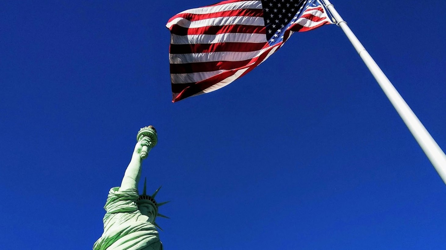 The American flag flying in the breeze on a clear day, with the Statue of Liberty's raised torch visible below against a deep blue sky