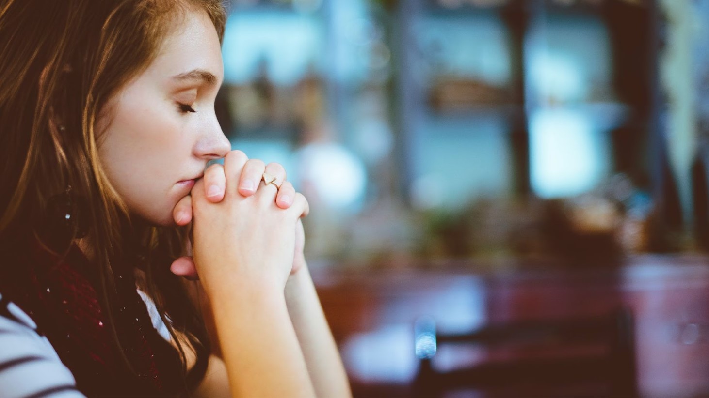 A young woman with closed eyes is captured in a moment of quiet contemplation or prayer, with her hands gently pressed together against her lips