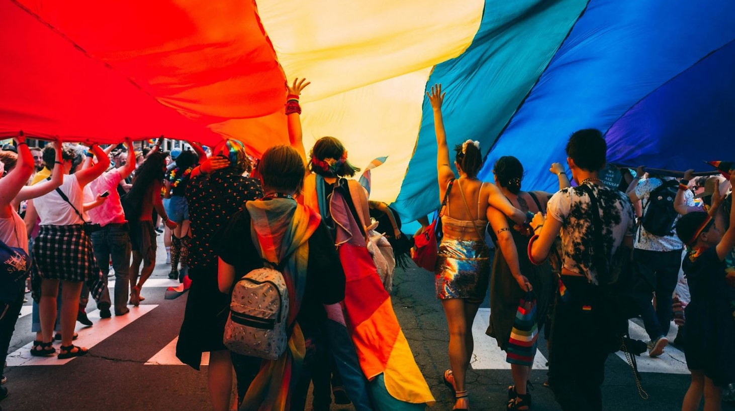 A vibrant scene at a pride parade, with a crowd of participants walking under a massive rainbow flag that they are holding up above them