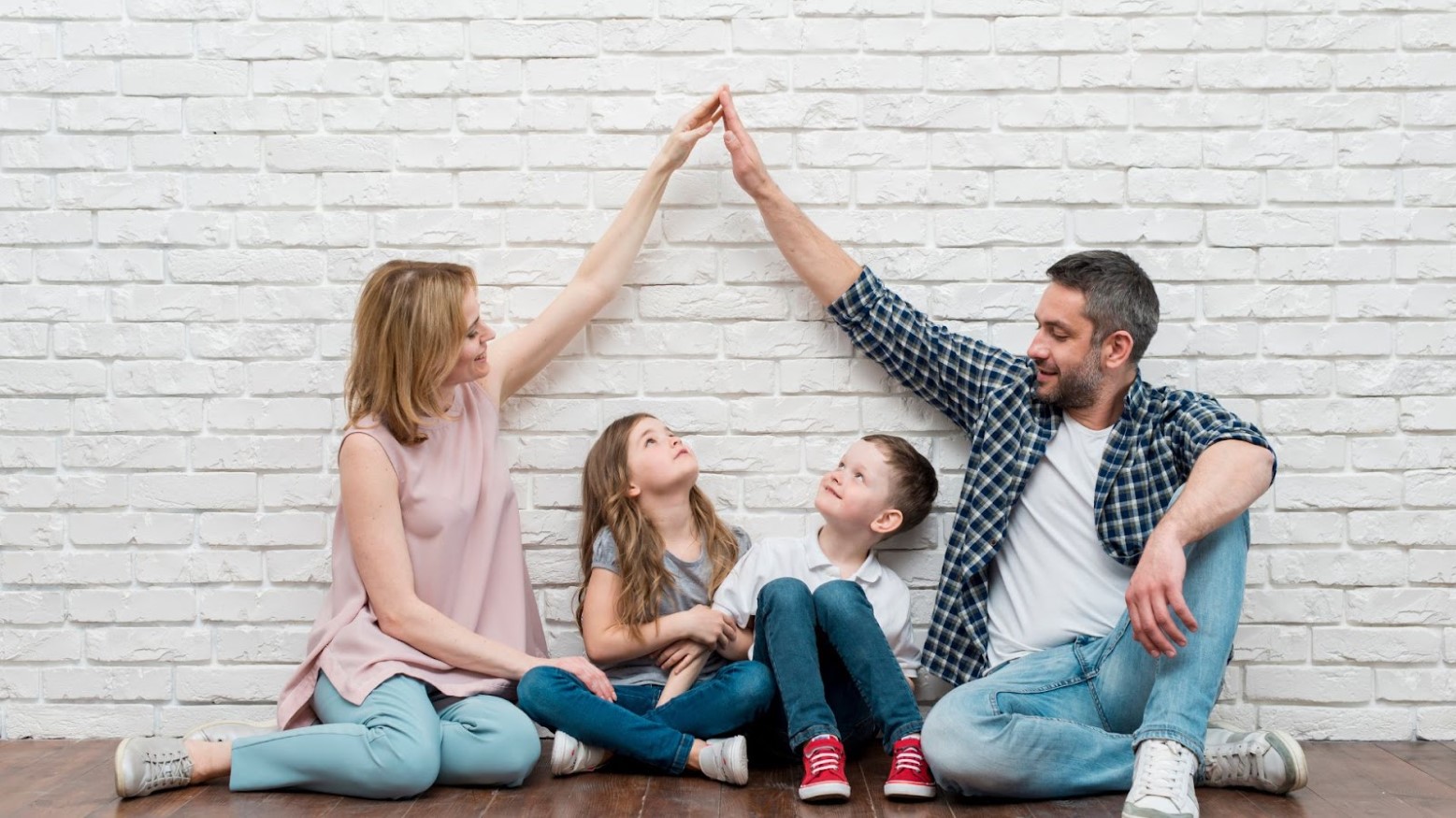 A young couple sits on the floor with their kids while holding their hands above the kids’ heads in an upside down V formation