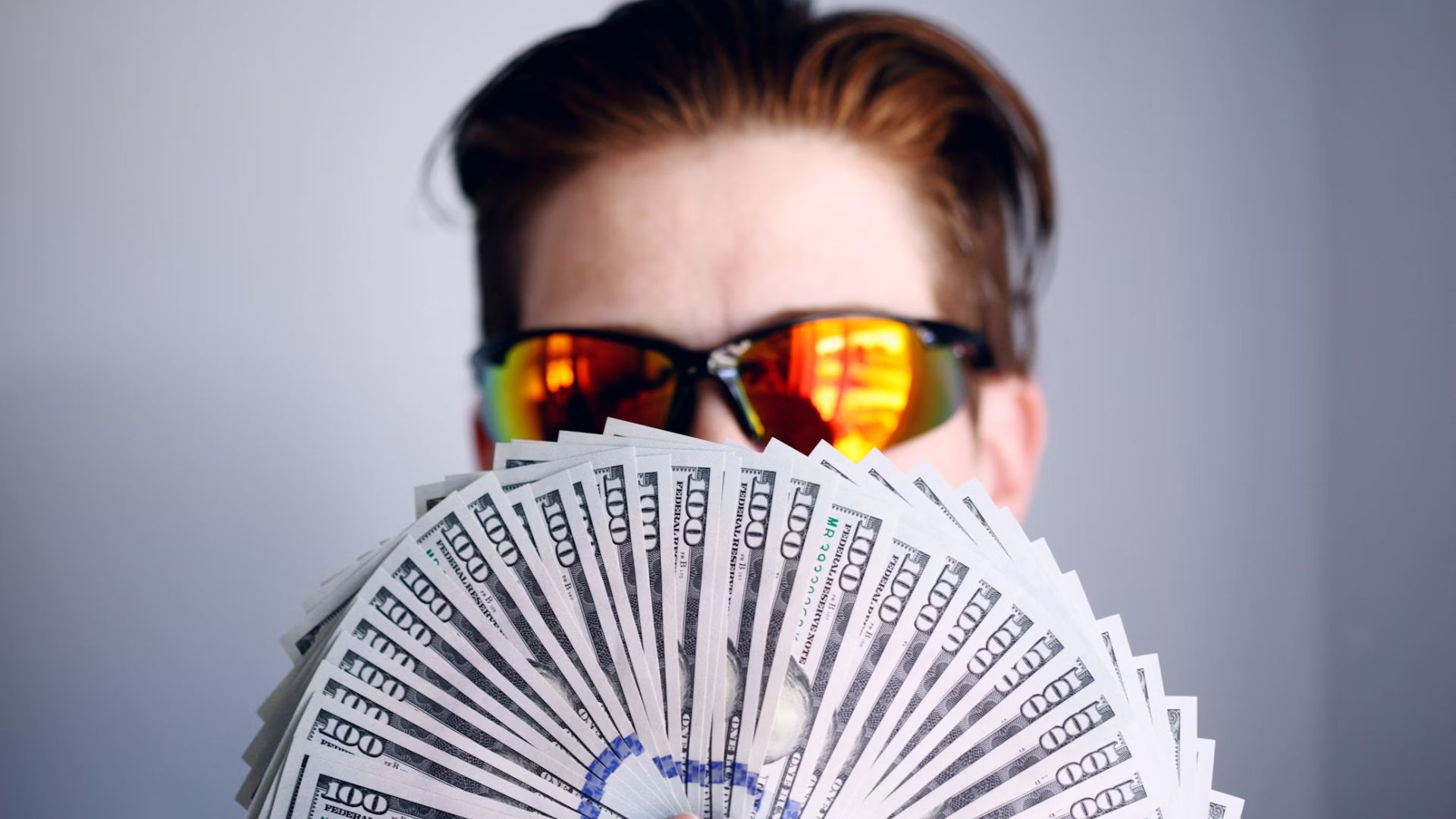 A man wearing orange sunglasses. He is holding many $100 notes fanned out