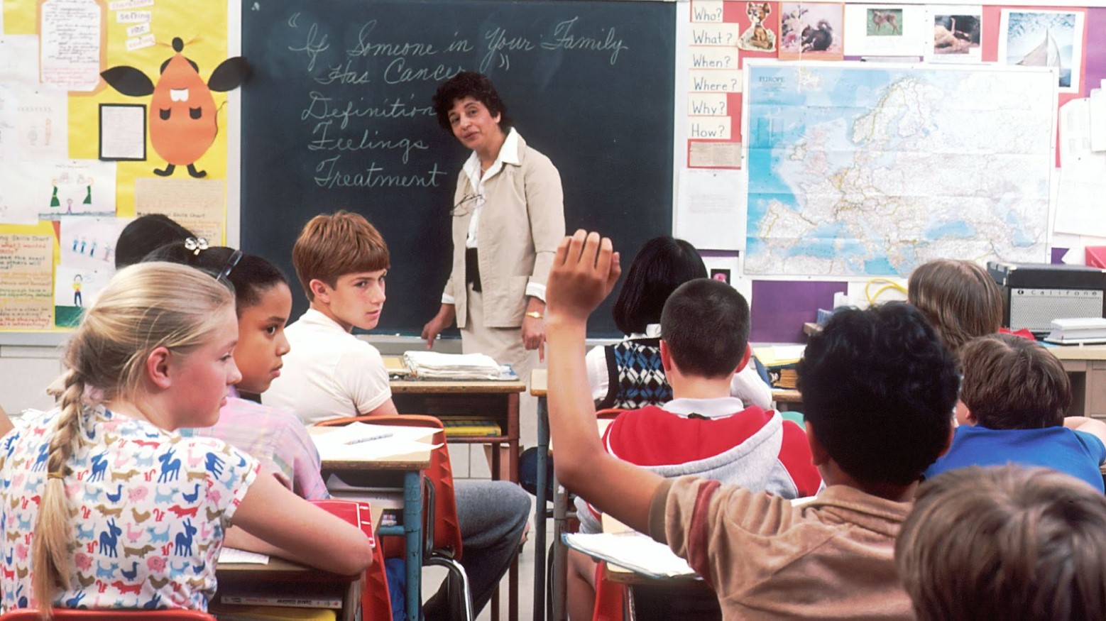 A teacher stands at a white board in a classroom during the 90s. Various students sit before her and one boy raises his hand